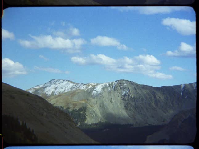 A vintage film shot captures a beautiful mountain landscape with snow-capped peaks under a blue sky dotted with clouds