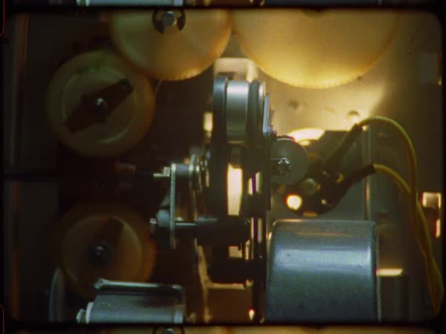 A close-up view reveals the intricate gears and mechanism of a vintage film projector in operation, glowing under the projection lamp