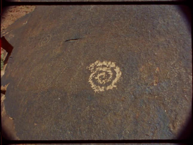 Vintage film footage with light leaks reveals an ancient spiral petroglyph etched onto a large boulder in a desert canyon