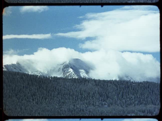 A vintage film shot captures a majestic, snow-covered mountain peak with clouds swirling around it, above a dense evergreen forest