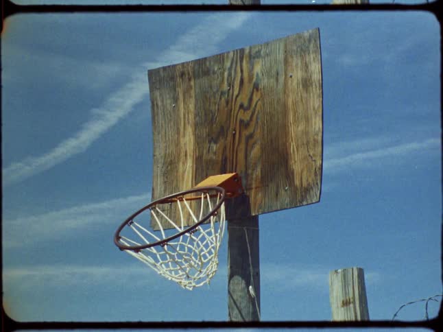 A rustic, homemade basketball hoop with a weathered wooden backboard stands against a clear blue sky in this vintage film footage