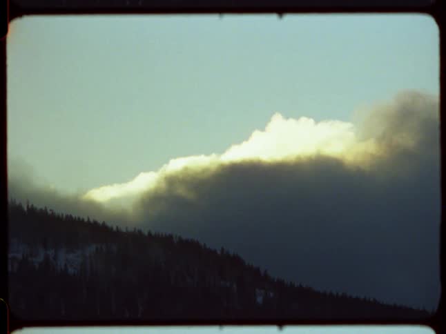 A dramatic, sunlit cloud drifts over a snowy, forested mountain in this vintage 8mm footage