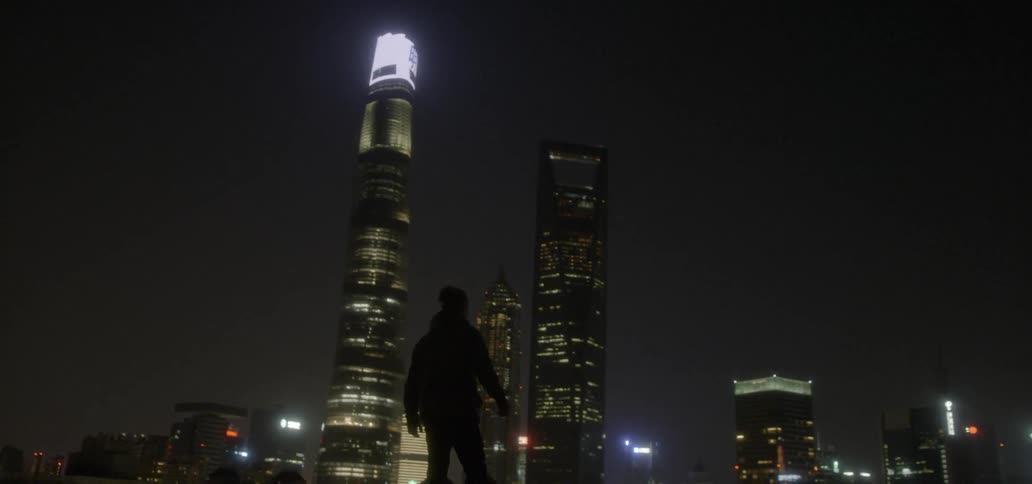 A man walks on a rooftop looking over a city at night