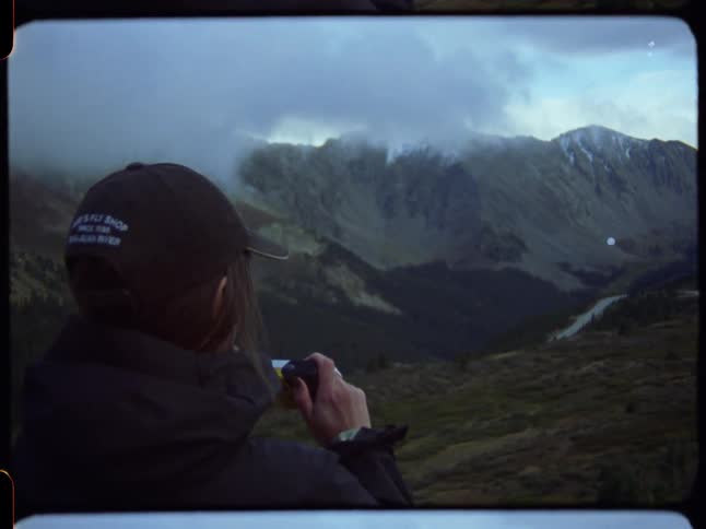 A person wearing a cap takes a photo of a cloudy, snow-capped mountain range on vintage-style film