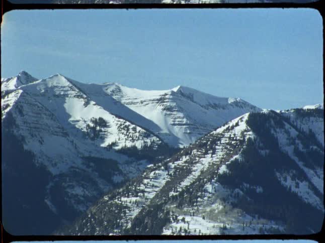 A vintage 8mm film captures a slow zoom into a majestic, snow-covered mountain range on a clear day