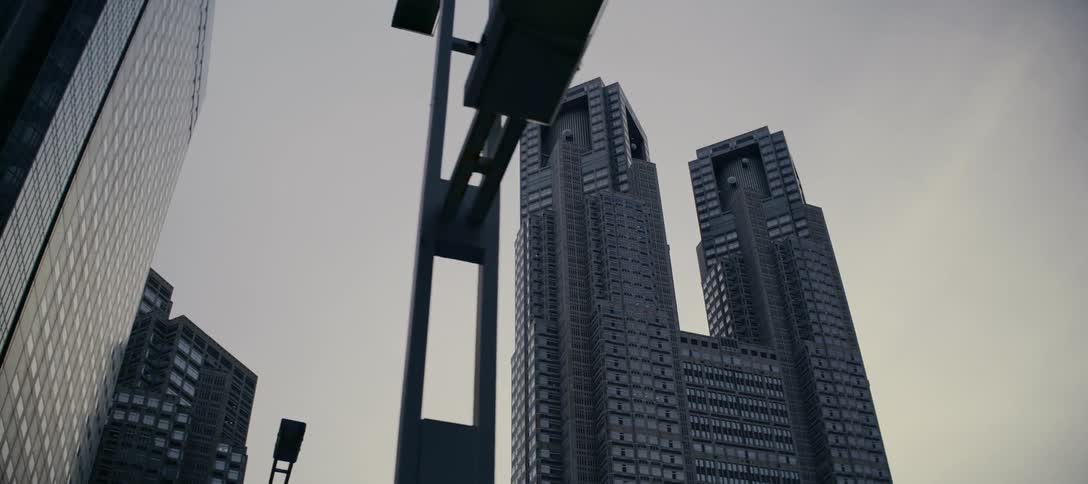 A slow, low-angle dolly shot reveals the imposing, futuristic architecture of the Tokyo Metropolitan Government Building against a grey sky