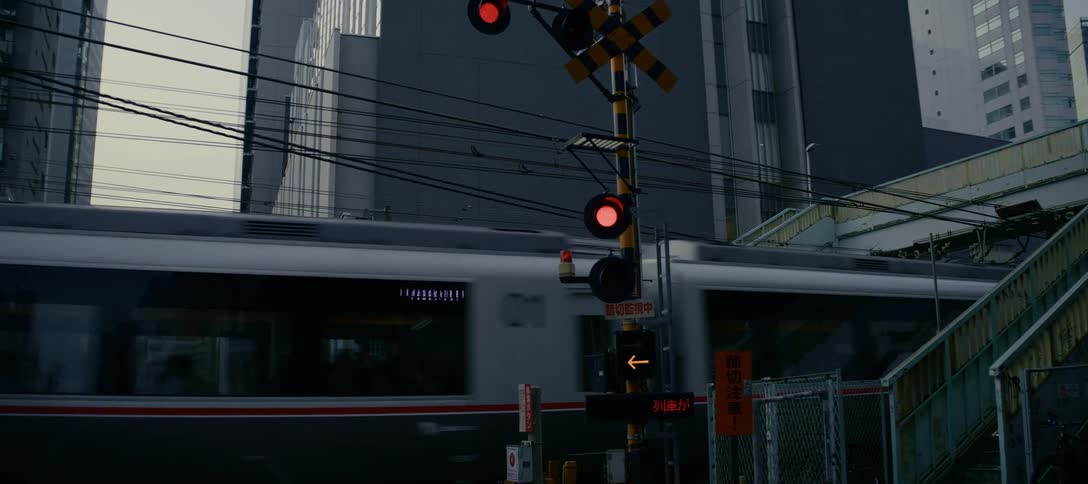 A slow zoom-in on a flashing railroad crossing signal as a passenger train speeds by in a Japanese city
