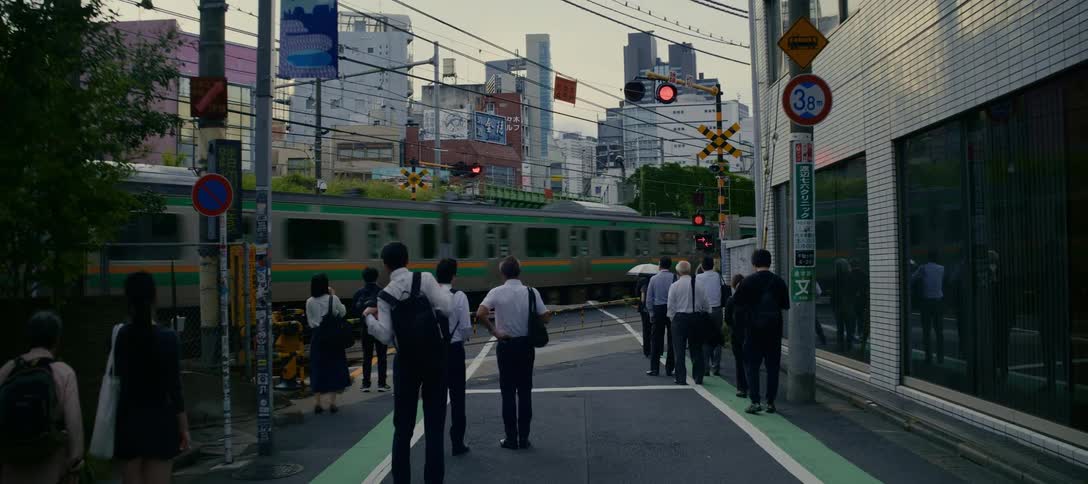 A slow zoom out reveals a group of Japanese commuters patiently waiting at a railroad crossing as a train passes by