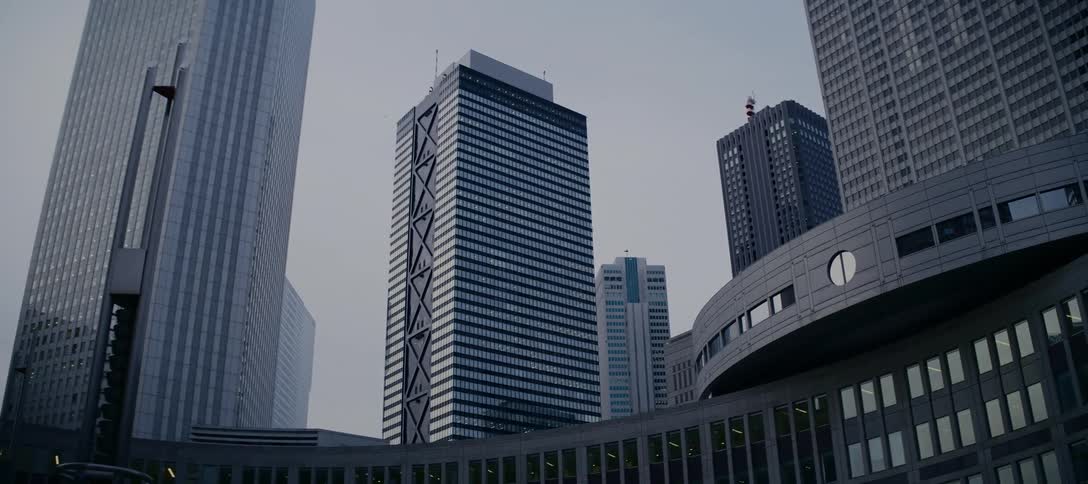A low-angle view of Tokyo's modern skyscrapers in the Shinjuku district, with an airplane flying high overhead against a grey sky