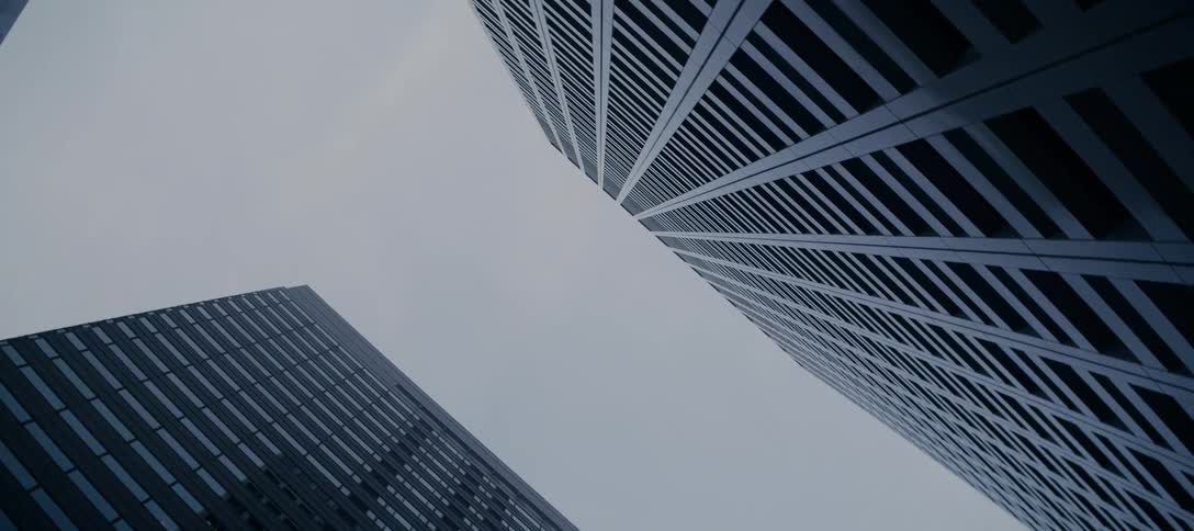 A slow, upward tilting shot looks up at two imposing modern skyscrapers against a bleak, overcast sky