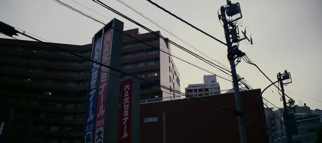 A slow pan reveals a quiet Japanese city street with dense buildings and tangled power lines under a gloomy, overcast sky