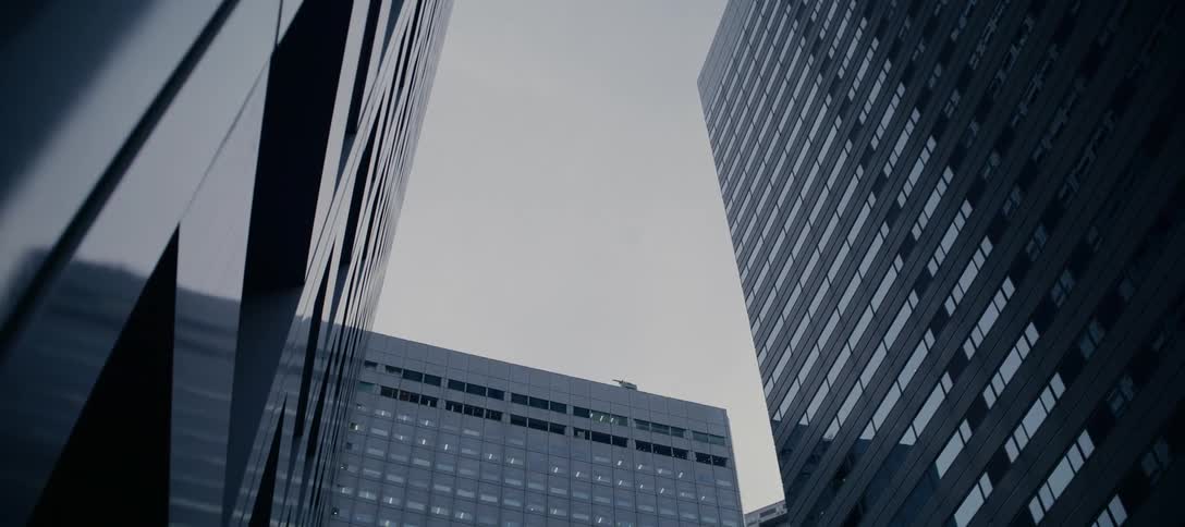A slow zoom-out shot from a low angle captures the imposing, modern architecture of towering skyscrapers against an overcast sky