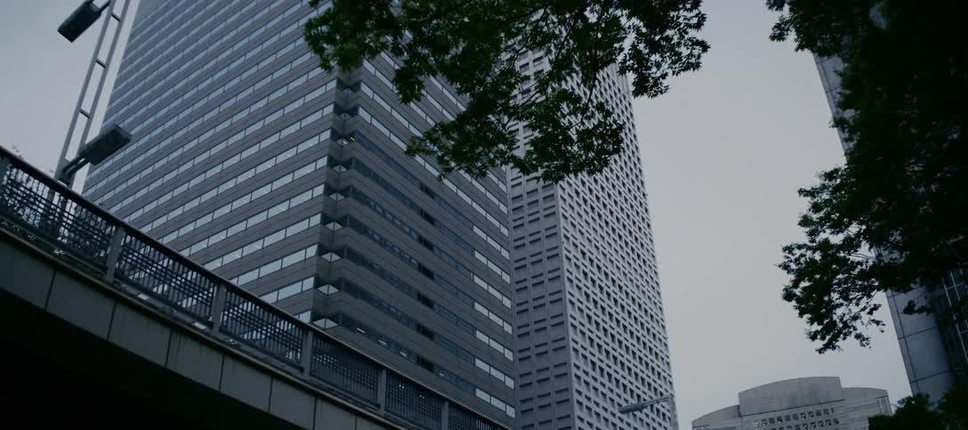 A low-angle view tilts down a modern, glass-facade skyscraper in an urban financial district on an overcast day