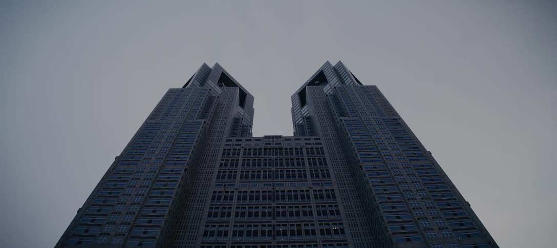 A low-angle view slowly tilts up the facade of a massive, symmetrical twin-tower skyscraper, revealing it against a flat gray sky