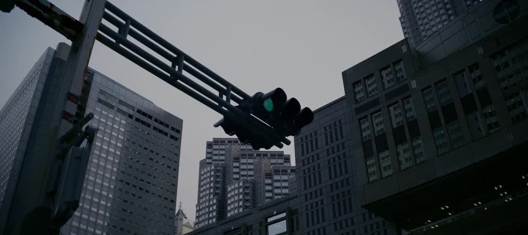 A green traffic light hangs against a backdrop of imposing grey skyscrapers under an overcast sky