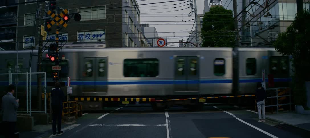 A silver commuter train speeds through a railroad crossing in a Japanese city as pedestrians wait for it to pass