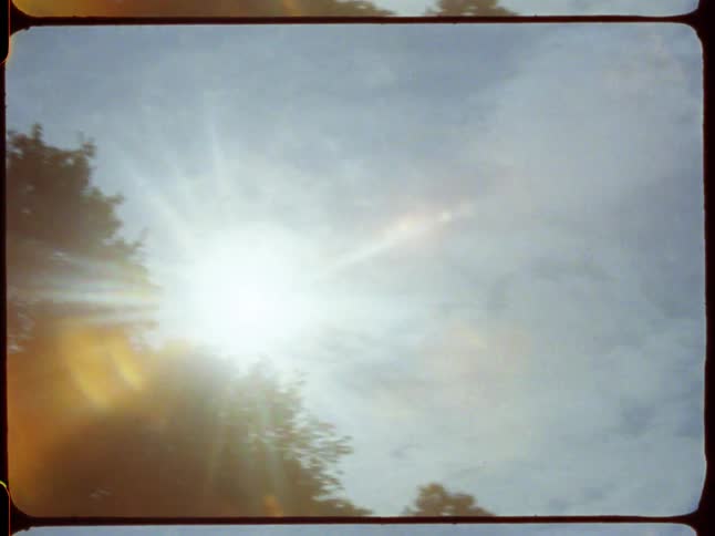 An 8mm film captures the sun shining through silhouetted trees, creating a beautiful lens flare against a cloudy blue sky