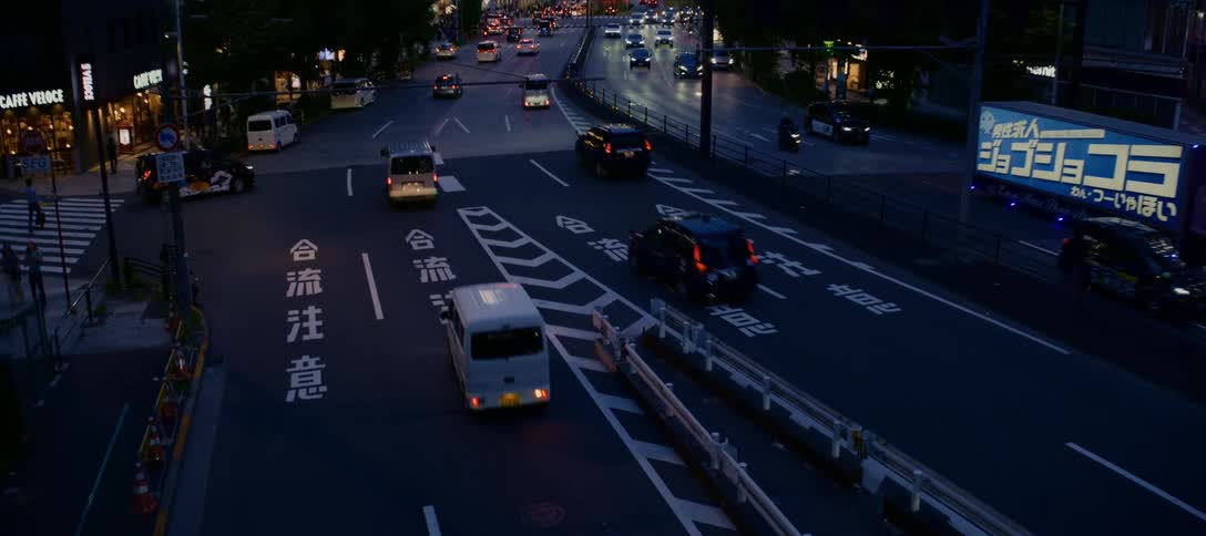 An elevated view captures the flow of evening traffic through a bustling Tokyo intersection, with vibrant neon signs illuminating the city skyline