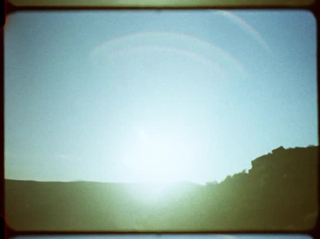 Vintage 8mm film captures a hazy sunrise over a grassy, hilly landscape, creating a prominent rainbow lens flare