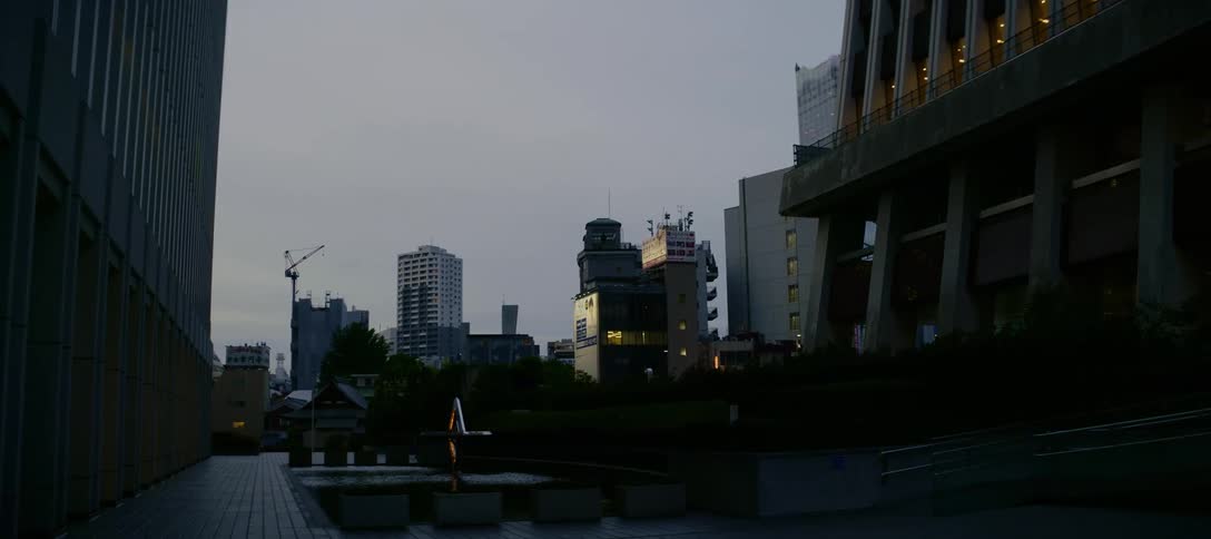 A low-angle shot tilts down from between two modern skyscrapers to reveal a quiet urban plaza at dusk