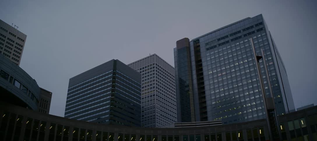 A slow low-angle pull-back shot reveals towering modern office buildings in a city's financial district under a gloomy, overcast sky