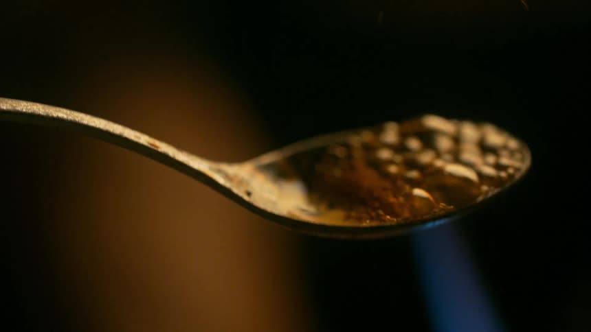 A close-up rack focus shot from an arm to a spoon of heroin bubbling as it's heated by a blue flame