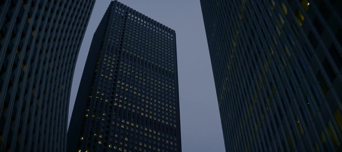 A slow, low-angle zoom-out reveals towering skyscrapers with lit-up office windows against the twilight sky