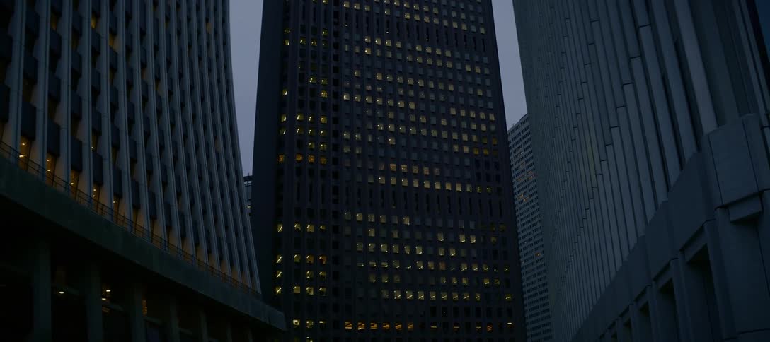 A quiet, low-angle tilt down reveals a modern skyscraper complex with glowing office windows at dusk