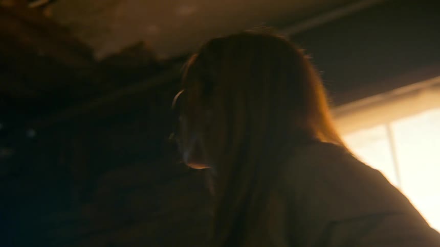 A distressed woman in a dark, abandoned room runs her hand through her hair and looks up in anguish
