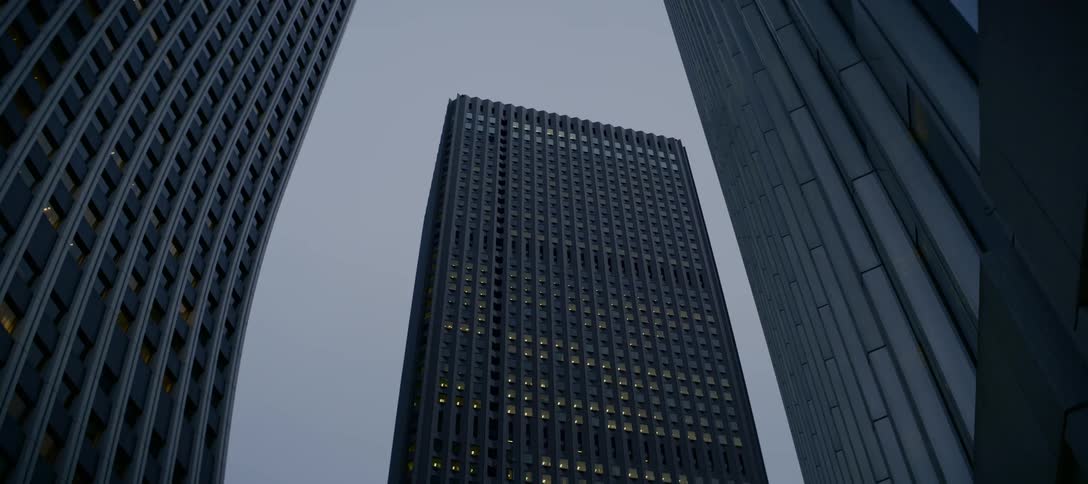 Slowly panning up a tall, dark skyscraper with lit windows in a modern city at twilight