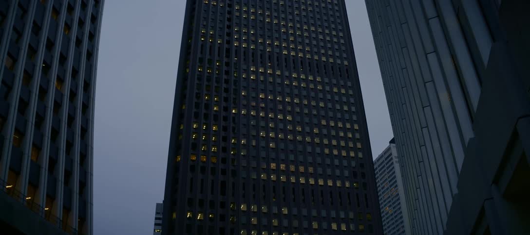 An imposing, modern skyscraper looms against an overcast sky at dusk as office lights glow from within