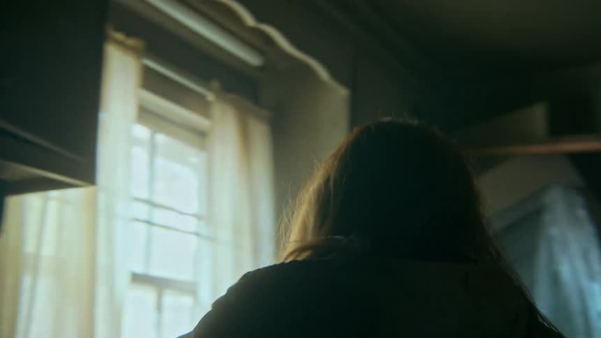 A distraught woman moves with frantic energy through a dark and cluttered kitchen, struggling to move a large appliance