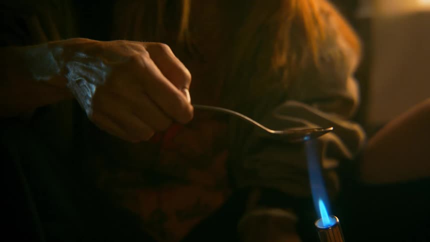 Close up on a distraught woman preparing heroin by heating a spoon with a blow torch in a dark, gritty room