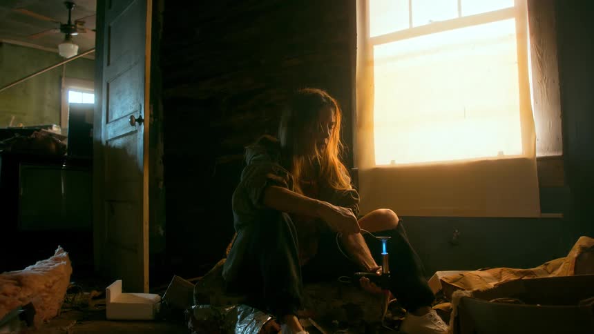 A woman prepares drugs with a lighter and spoon while sitting on the floor of a dark, messy, abandoned room