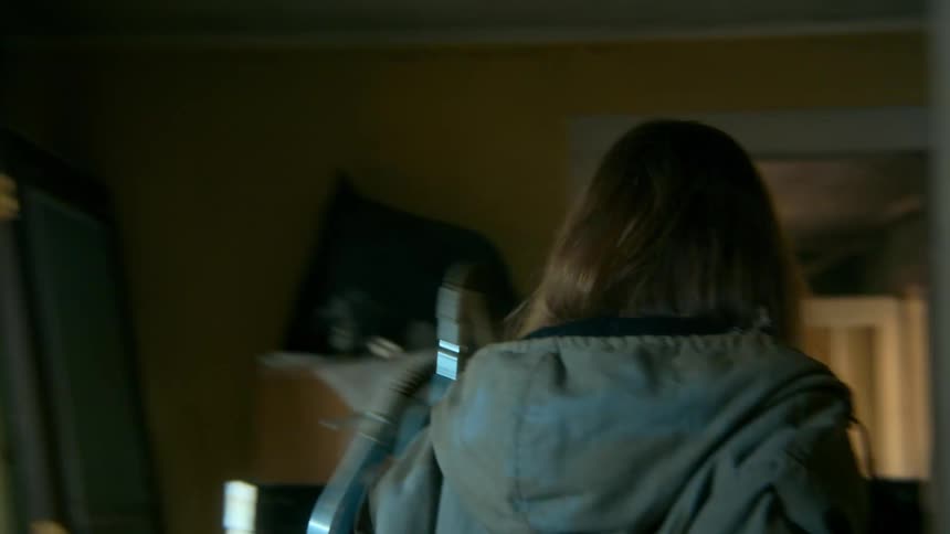 A disheveled and possibly injured woman wanders slowly and erratically through a dark and cluttered abandoned house