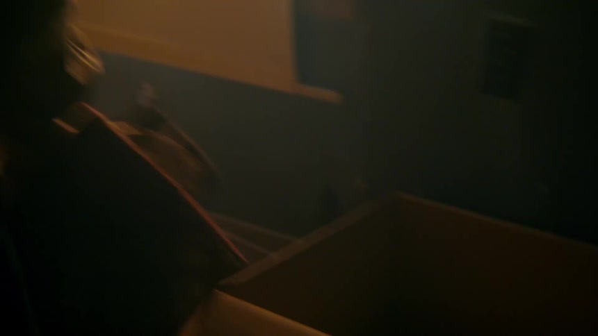 A distressed and disheveled woman frantically rummages through a cardboard box in a dark and dusty abandoned house