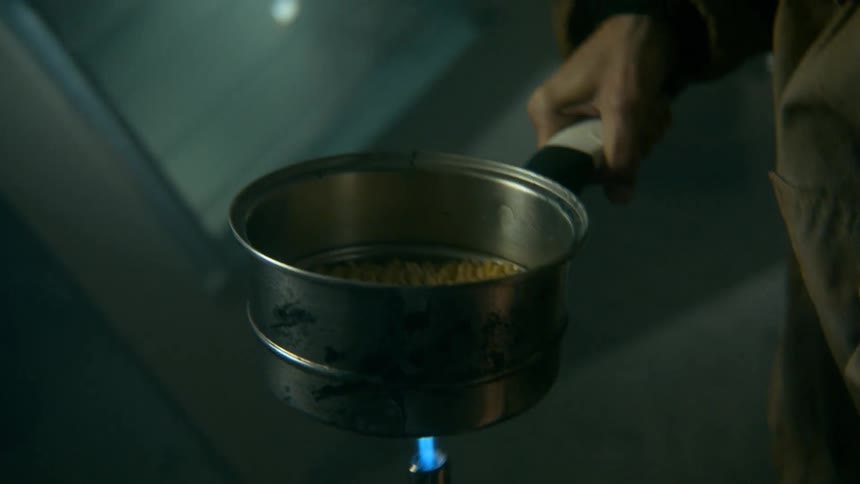 A woman in a dark, gritty environment uses a handheld torch to cook instant noodles in a pot