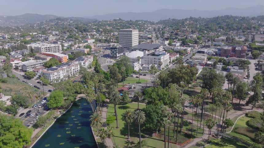 Slow aerial dolly shot over Echo Park Lake and its lush green surroundings towards the city of Los Angeles on a bright sunny day