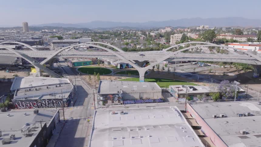 An aerial view pans left from the Sixth Street Viaduct bridge over a busy freeway to reveal the surrounding urban Los Angeles cityscape