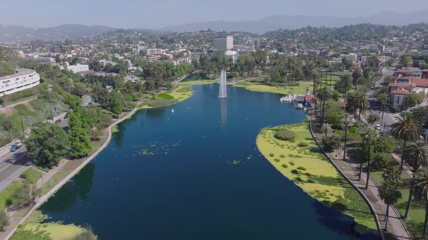 An aerial view slowly pushing in on the central fountain of Echo Park Lake, surrounded by the lush park and the sprawling cityscape of Los Angeles on a clear day