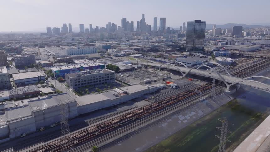 Expansive aerial view of a train traveling along the Los Angeles River, with the 6th Street Viaduct and the Downtown LA skyline visible on a clear day