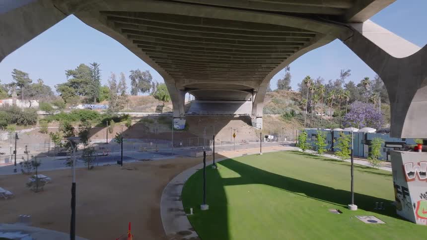 An aerial view slowly glides forward under a massive concrete freeway overpass, revealing a newly constructed but empty urban park below