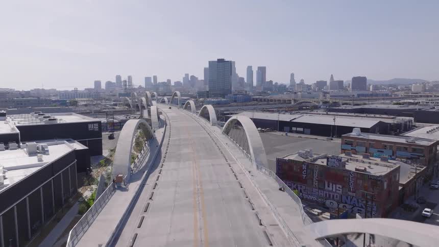 An aerial drone shot flies over the iconic 6th Street Viaduct bridge towards the downtown Los Angeles skyline on a clear sunny day