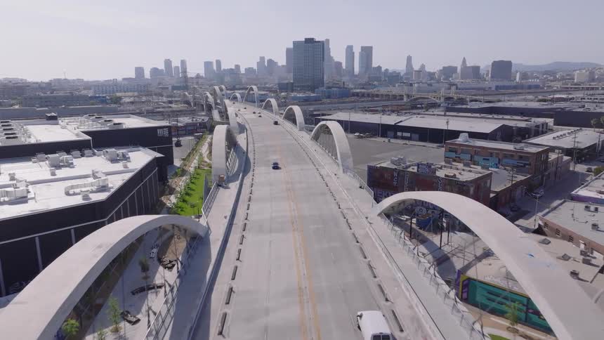 Aerial drone footage flies forward over the modern arches of the 6th Street Viaduct bridge towards the downtown Los Angeles skyline on a sunny day