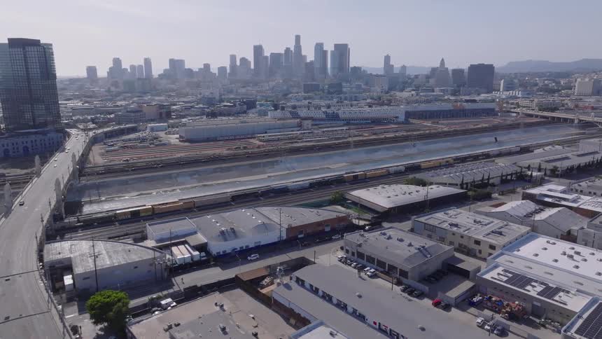 Slow aerial dolly shot over an industrial area with a railyard and bridge, moving towards the downtown Los Angeles skyline on a sunny day