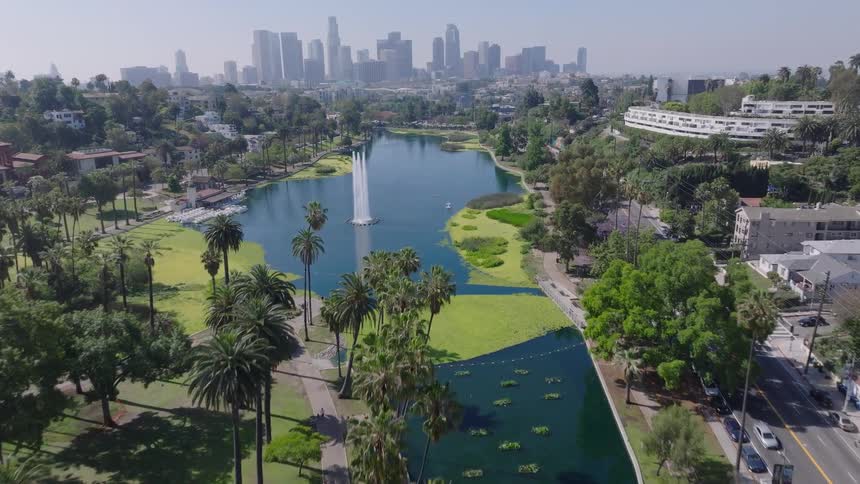 An aerial drone flies towards Echo Park Lake and its iconic fountain, with the downtown Los Angeles skyline visible on a clear day
