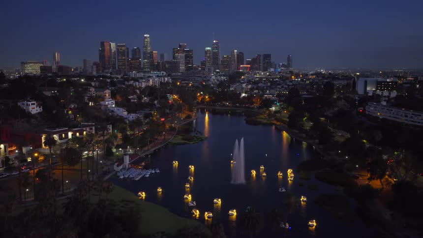 The iconic downtown Los Angeles skyline glows at night behind the illuminated fountain and swan pedal boats on Echo Park Lake