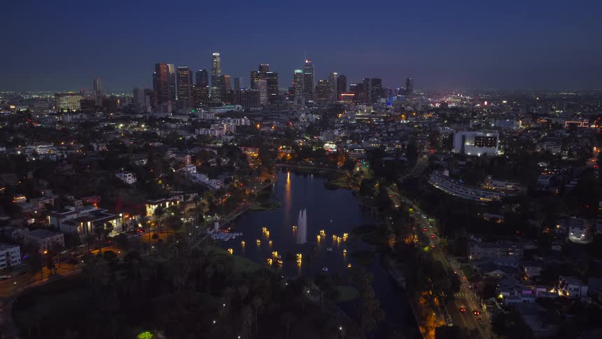 An aerial view of the Downtown Los Angeles skyline at night, with the illuminated fountain and boats of Echo Park Lake in the foreground