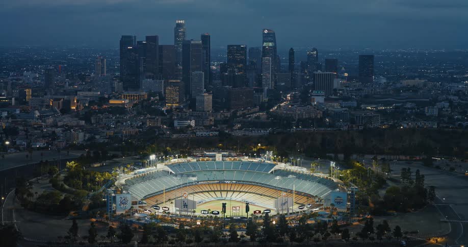 Slowly pushing in on an aerial shot of Dodger Stadium at dusk, functioning as a drive-in event venue with the glowing downtown Los Angeles skyline behind it