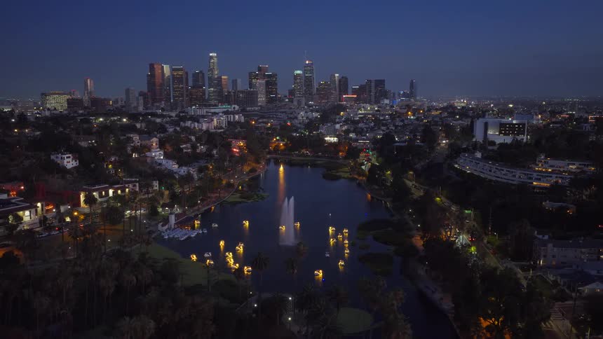 Ascending aerial view captures the beauty of Echo Park Lake at night, with its illuminated swan boats and fountain set against the vibrant Los Angeles downtown skyline